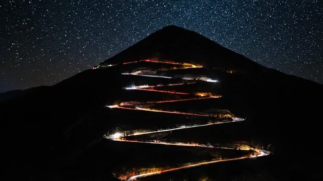 Vehicles ascend a steep, zigzag mountain road at night, their lights forming red and white trails against the dark silhouette, all beneath a dense field of stars.