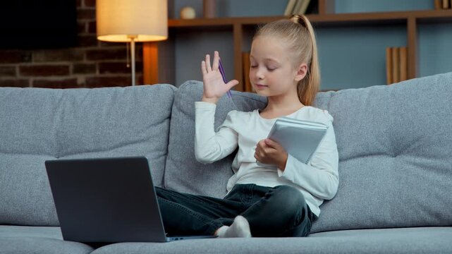Caucasian little cute school child girl kid e-learning raising hand arm ready to answer online tutor e-learning at home listening study lesson with computer and copybook on couch quarantine isolation