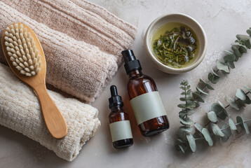 Two amber glass bottles with blank labels are placed on a marble surface alongside a wooden brush beige and cream towels a bowl of herbal infusion