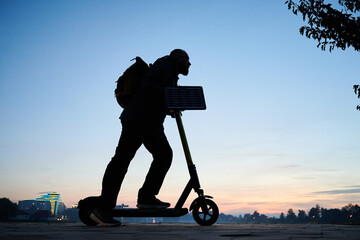 Man rides electric scooter while carrying solar panel. Integration of solar power as sustainable energy source for charging electric scooters, promoting eco-friendly urban transportation.