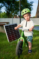 Young boy sits on green balance bike next to solar panel on grass. Kid wearing green helmet, looks at camera. Background features fence, trees, and bench. Concept of green energy generation.