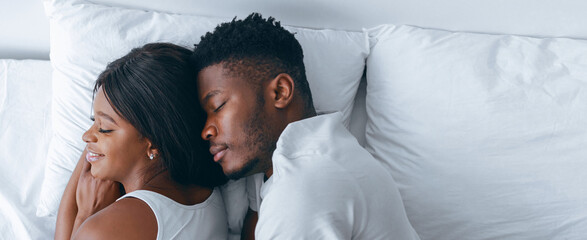 A couple is lying in bed together in a cozy space. The woman smiles while the man rests his head on her shoulder. Sunlight fills the room, creating a warm atmosphere.
