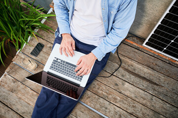 Top view of man works on laptop on lap and smartphone, sits on wooden deck. Solar panel nearby, emphasizing sustainable, modern eco-friendly workspace that combines technology and sustainability.