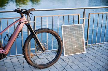 Pink electric mountain bike parked beside lakeside railing, connected to solar panel for charging. Bicycle's sleek design and rugged tires highlighted, with tranquil lake and golden light.