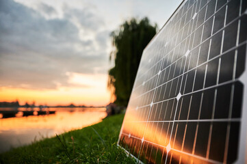 Solar panel illuminated by setting sun stands on grassy lakeside, with picturesque sunset and trees in background. Fusion of renewable energy technology with serene natural landscape.