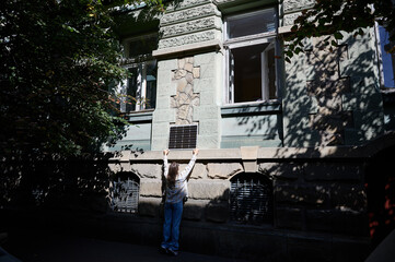Woman holding photovoltaic solar panel in front of historical building, wearing plaid shirt and sunglasses. Concept of integration of sustainable renewable energy sources into old architecture.