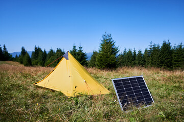 Yellow tent with solar panel set up in grassy field surrounded by dense evergreen trees under clear blue sky. Eco-friendly camping, integration of renewable energy in outdoor activities.