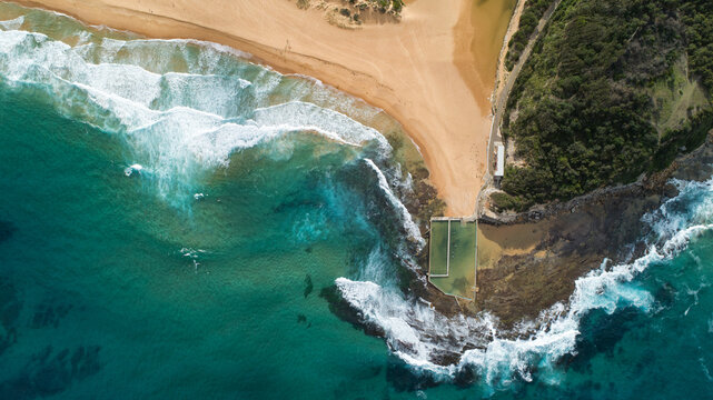 Aerial view of turquoise waves crashing against the golden sands of the beach and the rock pool at North Narrabeen, Sydney, New South Wales, Australia.