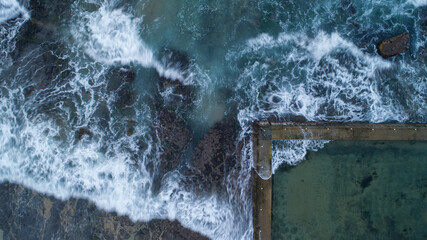 Aerial view of the powerful waves crashing against the rocks and the calm, contained ocean pool, contrasting textures and colors, Sydney, New South Wales, Australia.