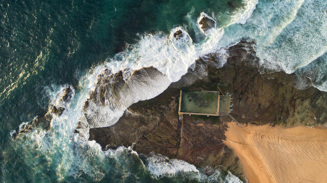 Aerial view of turquoise waves crashing against the rugged coastline, highlighting a man-made pool contrasting with the natural beauty of the shore, Sydney, New South Wales, Australia.