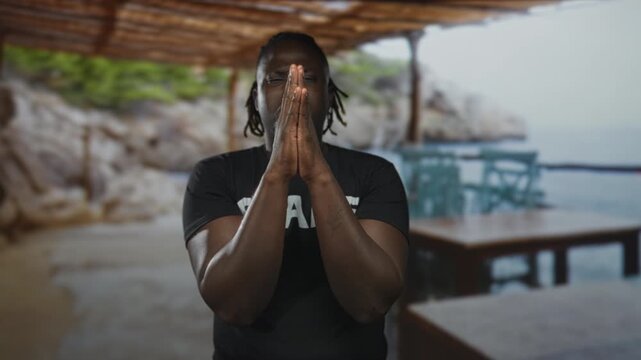 Man with hands pressed together praying at a seaside patio building under a timber pergola, eyes closed and focused; faith hope contemplation.