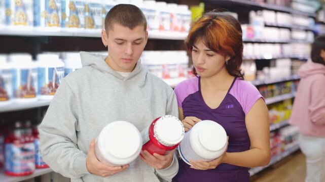 Guy and Latin girl consult about purchasing sports nutrition, enriched protein shake, carefully examines label on package. Shop visitor reads composition and characteristics of protein shake