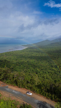 Aerial view of dense rainforest meeting the coastline near Cape Tribulation, with a road winding through the lush greenery, Cape Tribulation, Queensland, Australia.