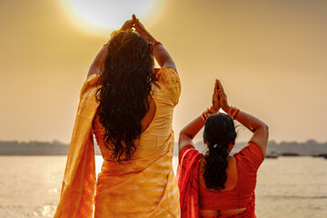 India. Uttar Pradesh state. Varanasi (Benares). Two Hindu women, dressed in yellow and red saris, stand facing the Ganges River, performing the prayer gesture (Namaskar or Surya Namaskar)