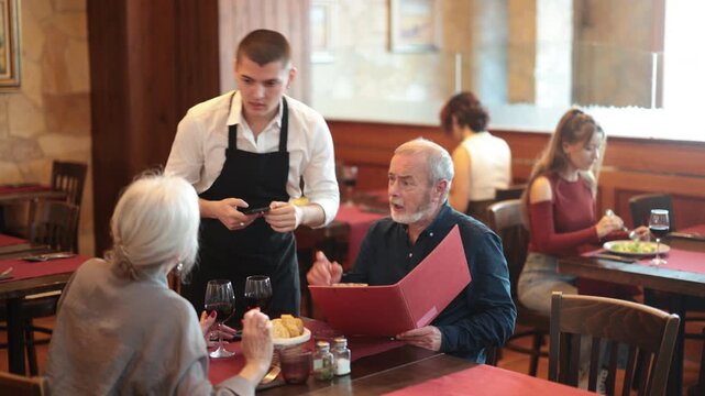 In restaurant hall, guy waiter brought order food to mature couple visitor, puts traditional Mediterrian salad of leafy greens, cheese on table. Waiter fetch and carry food for restaurant client