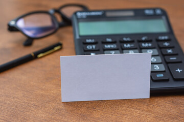 Empty white business card placed on a calculator over a wooden office desk. Professional corporate mockup for identity design, financial consulting, accounting services and branding presentations.