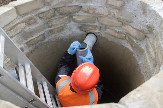 A worker wearing a protective helmet, safety gloves, and a high-visibility vest is installing a plastic sewer pipe inside a concrete sewage well at a private house. The image is taken from a top-down 