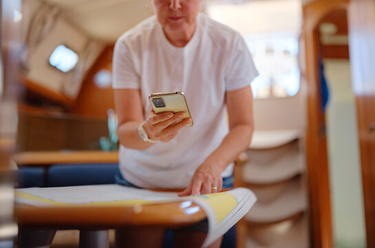 Mature woman sits inside yacht cabin analyzing paper sea chart for route planning. Navigation process involves calculating coordinates and path for upcoming sailing journey from Finike.