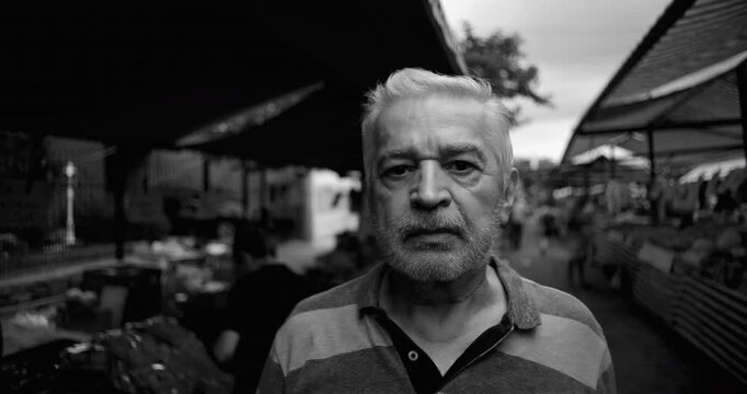 Older man paused at open air market with worried gaze meeting camera reflecting financial strain retirement insecurity and anxiety about food prices within everyday local commerce, black and white