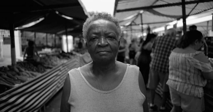 African American elderly woman standing at open air market with serious steady expression reflecting resilience economic pressure, daily commerce, black and white