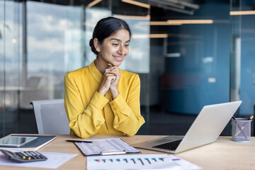 Smiling young Indian businesswoman sitting at office desk with documents and talking on video call...