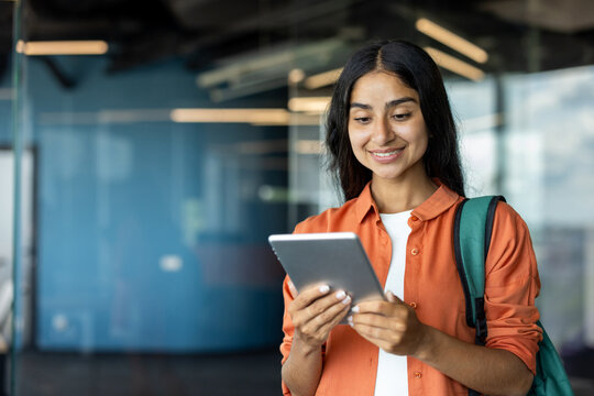 Young smiling Indian girl student with backpack standing indoors and using tablet