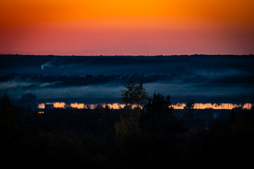 Obraz premium A serene sunset landscape with warm orange sky reflecting on a calm river, layered with misty fog over a rural village and forest horizon, creating a peaceful and atmospheric evening scene.