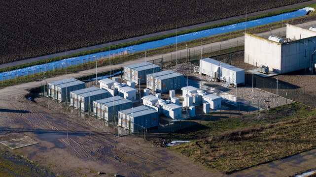 Aerial view of a cluster of shipping containers and industrial buildings, bordered by a narrow canal and fields, creates a stark contrast between man-made structures and the natural landscape.