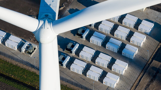 Aerial view of a stark white wind turbine standing tall beside rows of neatly arranged white containers, creating a geometric contrast, Eemshaven, Netherlands.
