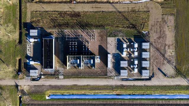 Aerial view of an electrical substation, a concrete island amidst fields of brown, with buildings casting stark shadows, Netherlands.