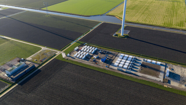 Dronten, Netherlands - 14 January 2026: Aerial view of a stark contrast between dark, tilled earth and vibrant green fields, a wind turbine standing tall amongst the energy storage units.