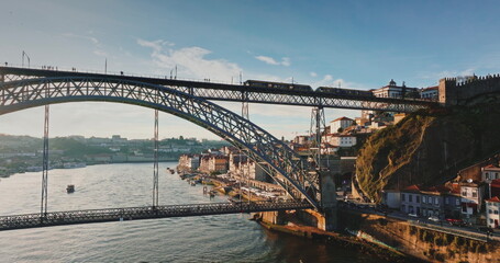 Portugal: Dom Luis I Bridge spans Douro River in Porto, with commuter train on upper deck and traffic below, framed by historic riverfront buildings at golden sunset hour. Aerial view drone