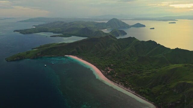 Drone footage at sunrise over Padar Island in Komodo National Park, Indonesia, revealing Pink Beach, lush green hills, turquoise water and calm tropical sea during soft golden morning light in 4K.