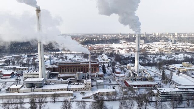 Drone aerial of Vilnius combined heat and power plant: twin chimneys vent winter steam above snowy rooftops, forest edge and city skyline.