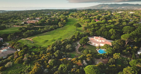 Portugal, Lisbon: Aerial view golf course in Cascais with lush green meadow ad trees landscape, luxury villas houses, mountain and ocean in background. Beautiful sunset scene. Drone flight