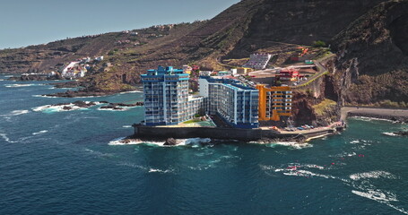 Tenerife: Modern residential buildings and resorts on rugged coastline of Atlantic Ocean, waves breaking on rocky shore and winding road ascending the volcanic mountain landscape. Aerial view panorama