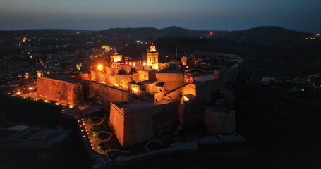 Malta, Comino island: Warm lights illuminating the Cittadella in Gozo at twilight, creating magical atmosphere reflecting Maltese history and old architecture at night. Aerial drone flight