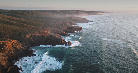 Portugal, Lisbon: Dramatic rugged cliffs and verdant hills lining the Atlantic Ocean coastline, with golden hour sunlight illuminating the landscape, waves crash against the shore. Aerial drone flight