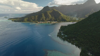 Moorea island aerial view with lush green mountains tumbling into a clear blue lagoon, small coastal village along a pristine shoreline under a partly cloudy tropical sky