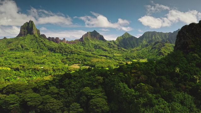 Moorea island displaying dramatic volcanic peaks and valleys covered in dense, vibrant green tropical jungle with a bright blue sky and scattered white clouds overhead