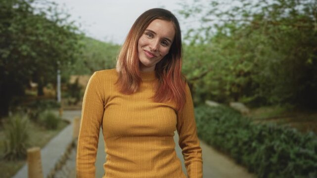 Woman smiling with tilted head in mustard turtleneck sweater on a tree lined street path with bollards and lush greenery; contentment.