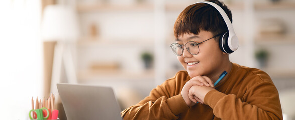 A young boy with glasses and headphones sits at a desk working on a laptop. He is smiling and holding a pen, engaged in an online class. Pens and a lamp are on the desk.