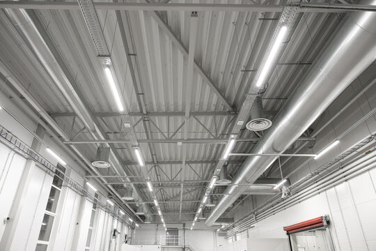 High ceiling of a modern industrial facility featuring exposed metal beams, suspended LED lighting, and large ventilation ducts. Bright, clean environment with copy space