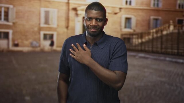 Man with hand on chest standing in cobbled courtyard in front of historic building, calm expression and slight smile, gesture of pledge and thanks; gratitude reflection.