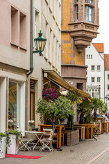Obraz premium A section of an old street where wooden cafe tables and palm trees stand near houses. A square lantern hangs on the wall. The street is in Hanover, Germany