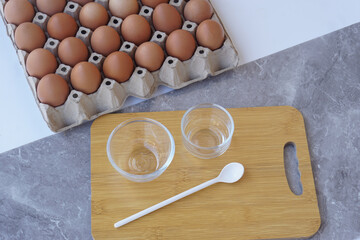 Top view of fresh brown chicken eggs in paper carton with glass bowls and a white spoon on  wooden cutting board. Concept for cooking, baking, food ingredients, and healthy homemade meals preparation