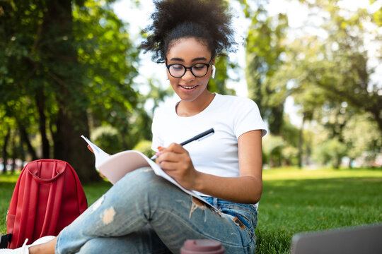 Happy African American Student Girl Learning Using Laptop And Writing Taking Notes Wearing Earbuds Sitting In Park Outdoor. Studentship Leisure And Lifestyle. Online Education Concept