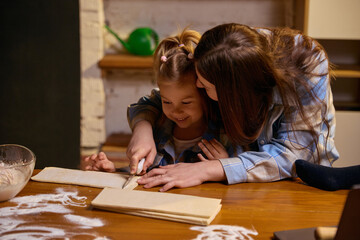 Mother teaching young daughter to cut dough at kitchen table at home. Concept of family cooking,...