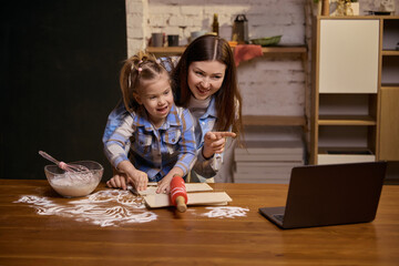 Fototapeta premium Mother and daughter smiling while preparing dough at home table. Concept of positive family routine, home cooking, parent child bonding, lifestyle photography, everyday domestic activity.