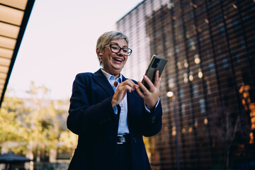 Happy senior woman using smartphone outdoors with wireless earbuds, expressing joy and connection during digital communication, showcasing emotional tech engagement.